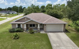 A single-story beige house with a two-car garage, gable roof, and front porch sits on a grassy yard with shrubs and trees around—an inviting setting perfect for roofing contractor Southwest Florida services under the partly cloudy sky.